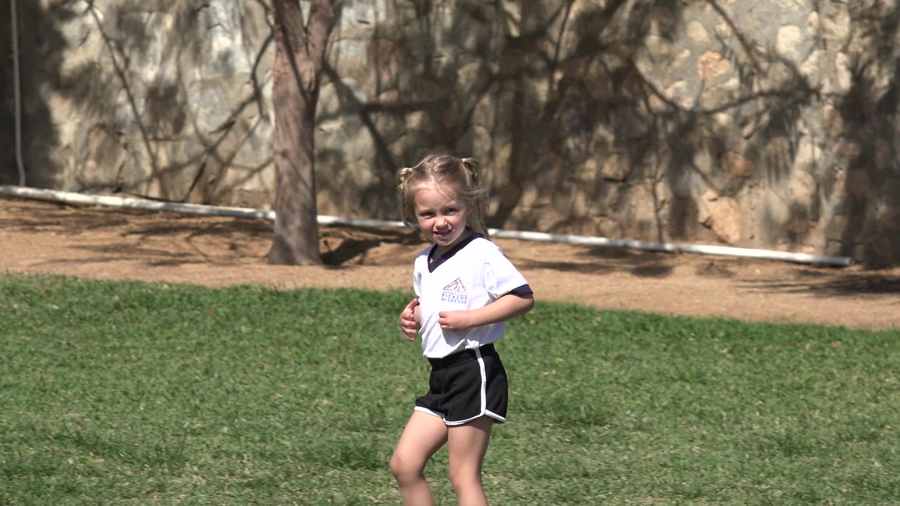 The young blond girl remains near the center of the frame on green grass in Los Cabos, Mexico, wearing a white shirt with dark trim and black shorts.