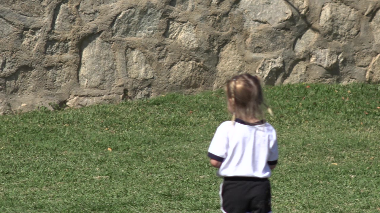 A young girl in a white shirt and black shorts stands or walks away from the camera on grass near a stone wall in Los Cabos, Mexico.