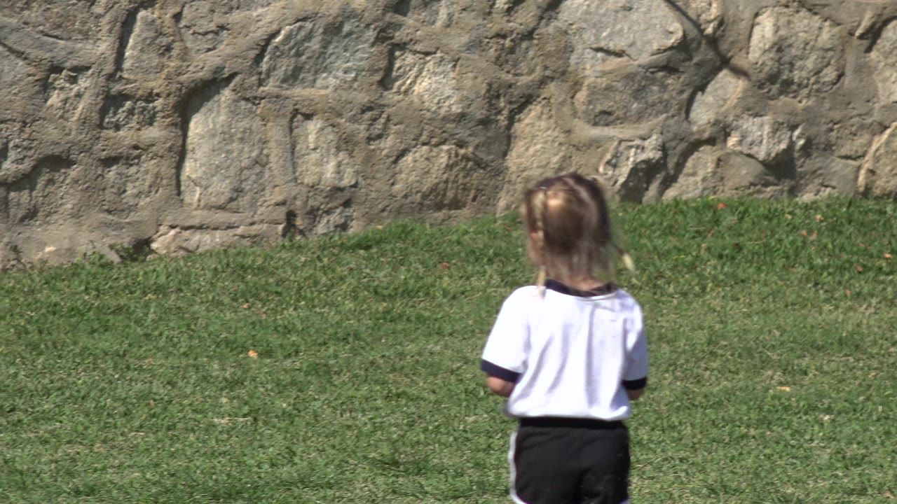 The girl remains centered with her back to camera on sunlit grass, with the rough stone wall filling the background in Los Cabos, Mexico.