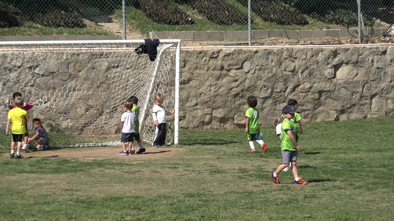 Children in green jerseys, white shirts, and a yellow shirt stand by the goalmouth in Los Cabos, Mexico, while one child walks across the grass in front.