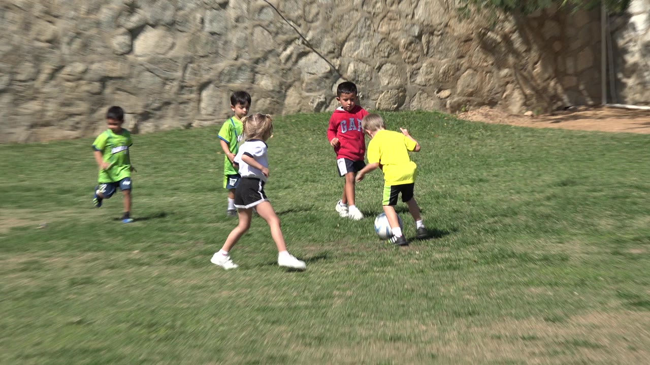 A blonde girl in a white jersey moves toward boys in bright shirts near a soccer ball on the grass in Los Cabos, Mexico.