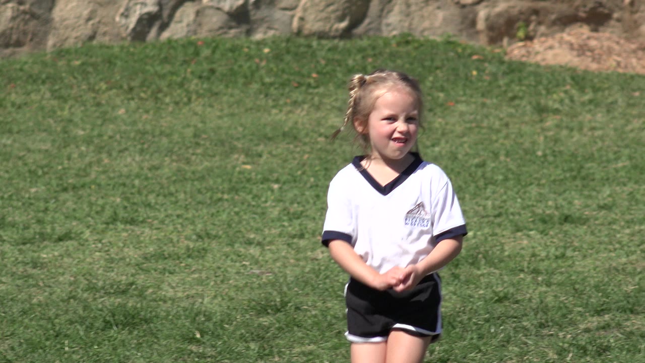 A young girl in a white sports-style shirt stands on green grass in Los Cabos, Mexico, with rocks in the background at the start of the clip.