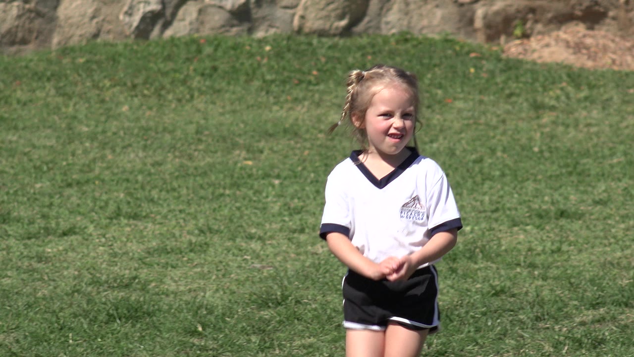 The girl remains centered on the grass in Los Cabos, Mexico, with her hands together and a small smile visible in bright daylight.