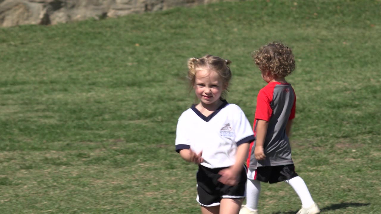 A young girl in a white soccer shirt and black shorts stands on a sunlit grass field in Los Cabos, Mexico, with another child in a red and gray jersey behind her.