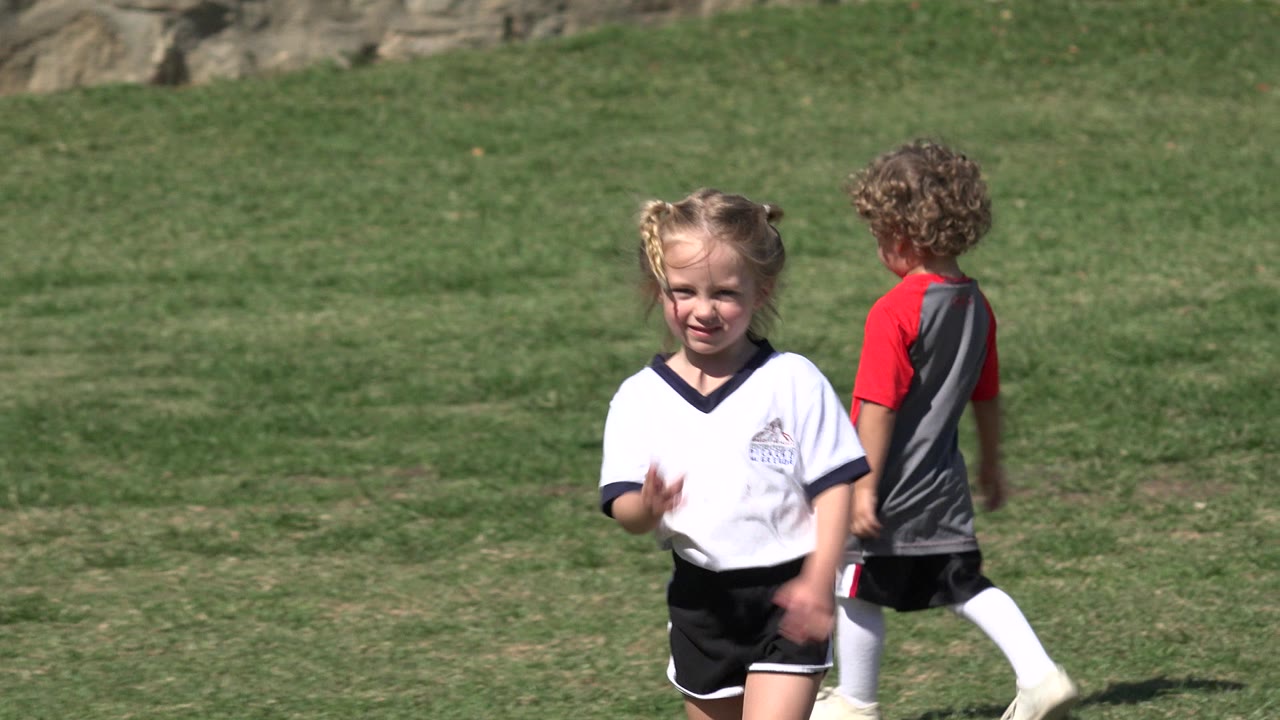 The girl faces the camera on the Los Cabos grass field while a child in a red and gray jersey stands nearby and another child in green runs farther back.