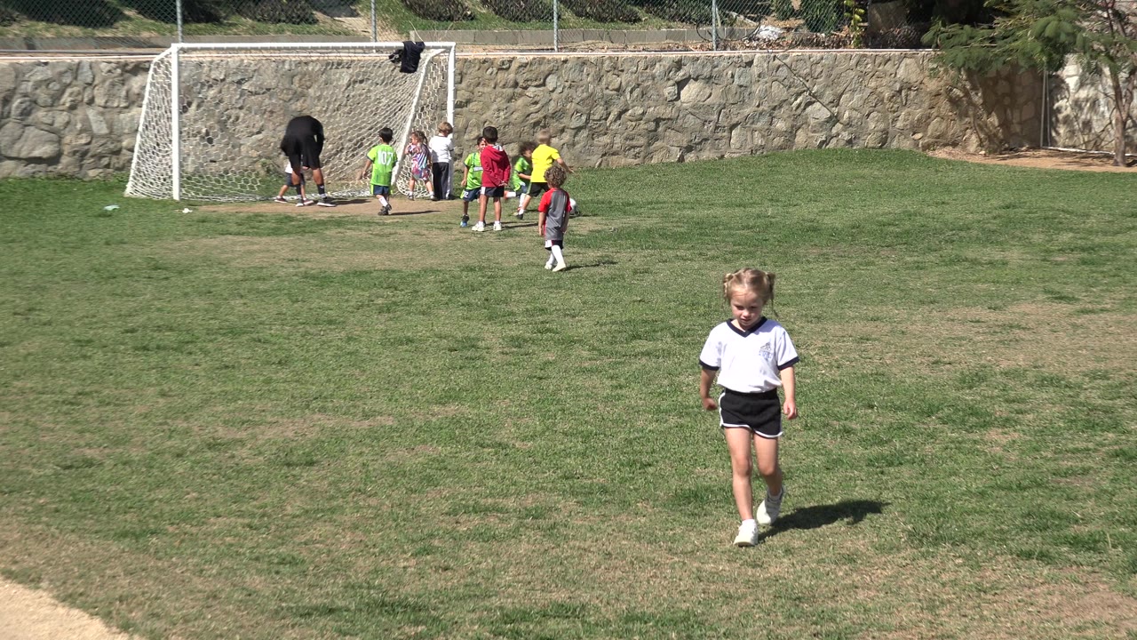 Young girl walking on a soccer field with children near a goal behind her.