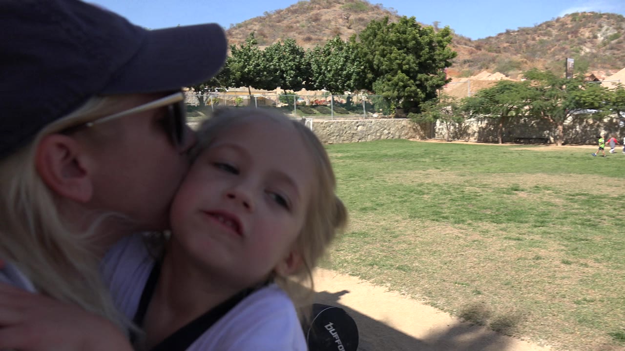 In Los Cabos, Mexico, the young girl faces the camera while leaning into the woman under shade, with open grass, small distant players, trees, and hills behind them.