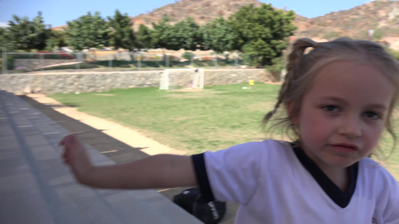 The blonde girl remains close in the foreground in Los Cabos, Mexico, with one arm extended while the soccer field and goal stay visible behind her.