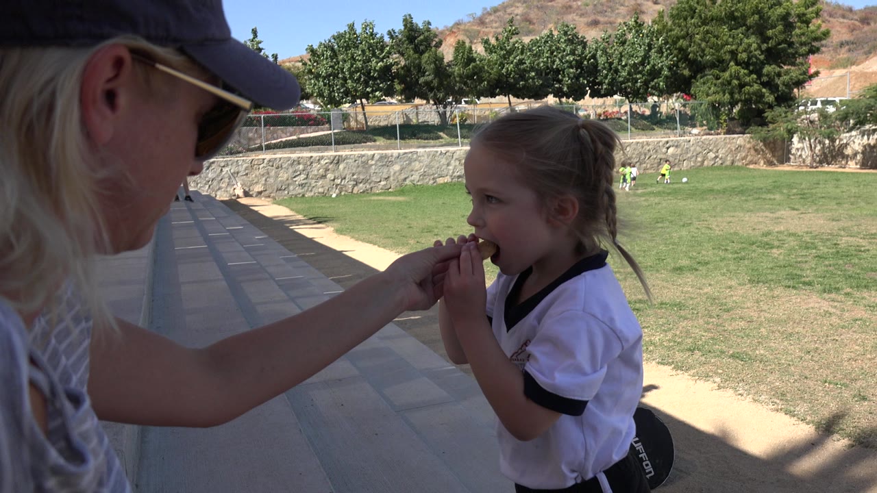 In Los Cabos, Mexico, a woman in sunglasses and a cap reaches from the shaded sideline to feed a small girl in a white sports shirt, with a green field and trees behind them.