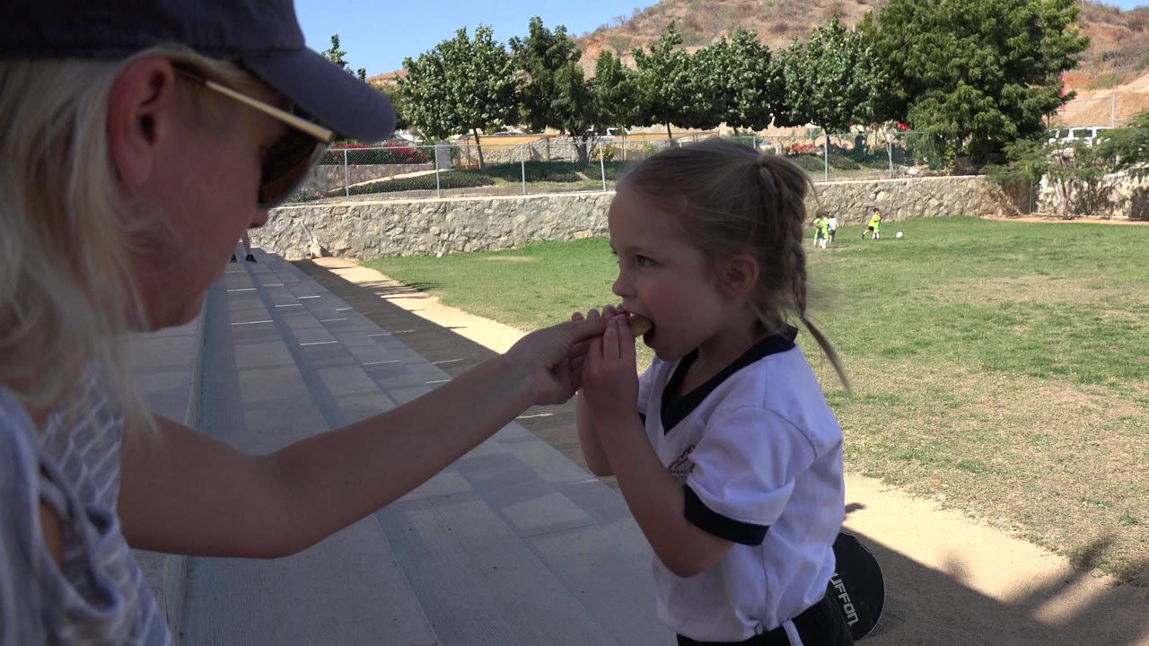 The girl continues eating from the adult's hand on the shaded edge of the Los Cabos sports field, with distant players and a tree line in the bright background.