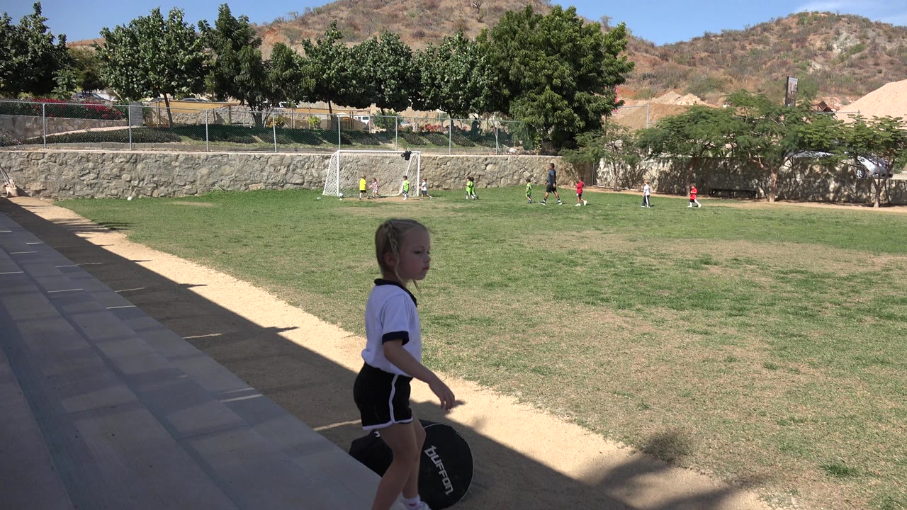 Young girl walking beside tan bleachers near a soccer field.