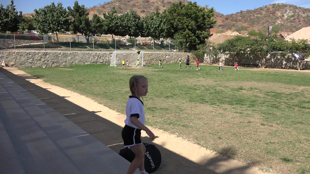 The girl continues across the lower frame in Los Cabos, Mexico, while bright grass, a white soccer goal, trees, and dry hills fill the background.