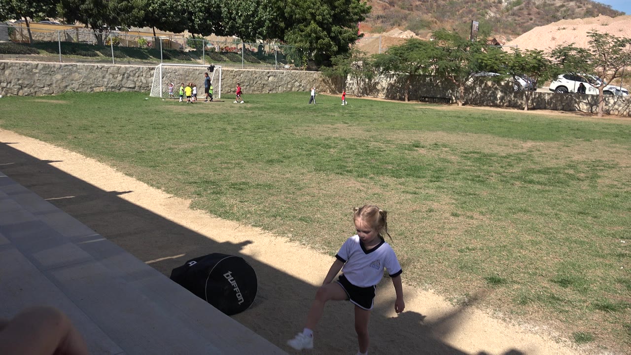 One second in, the girl remains near the lower frame beside the shaded walkway, with the grass field, goal, and small groups of children visible in Los Cabos behind her.