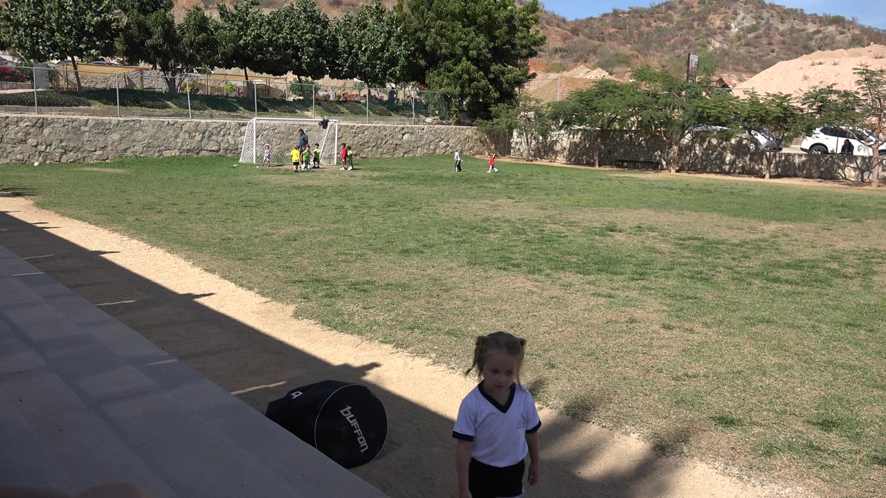 Girl at lower right with a wide sunlit soccer field behind her.