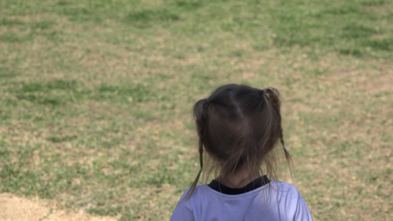 At the start of the Los Cabos clip, a young blonde girl is framed from behind with her head near the lower center of a sunlit grassy field.