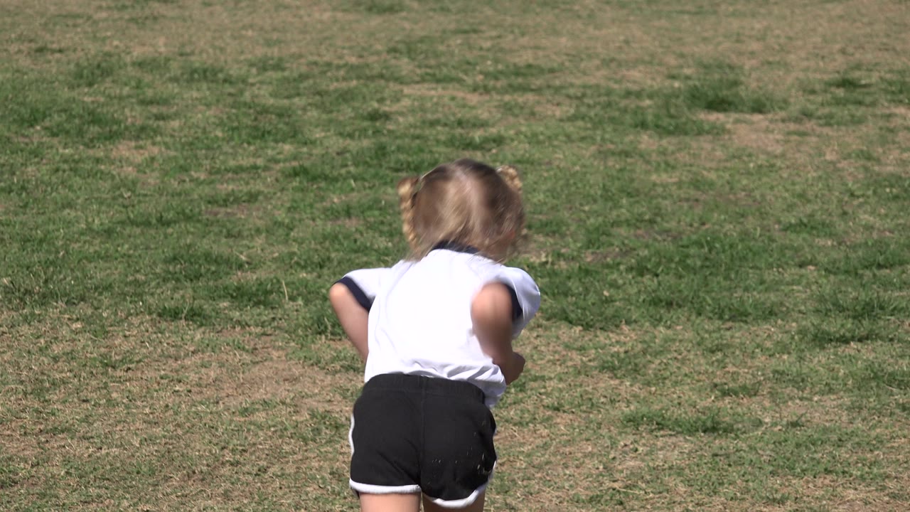At 0:01, the child continues running away in the center foreground, with the sunlit grass field filling the frame in Los Cabos, Mexico.