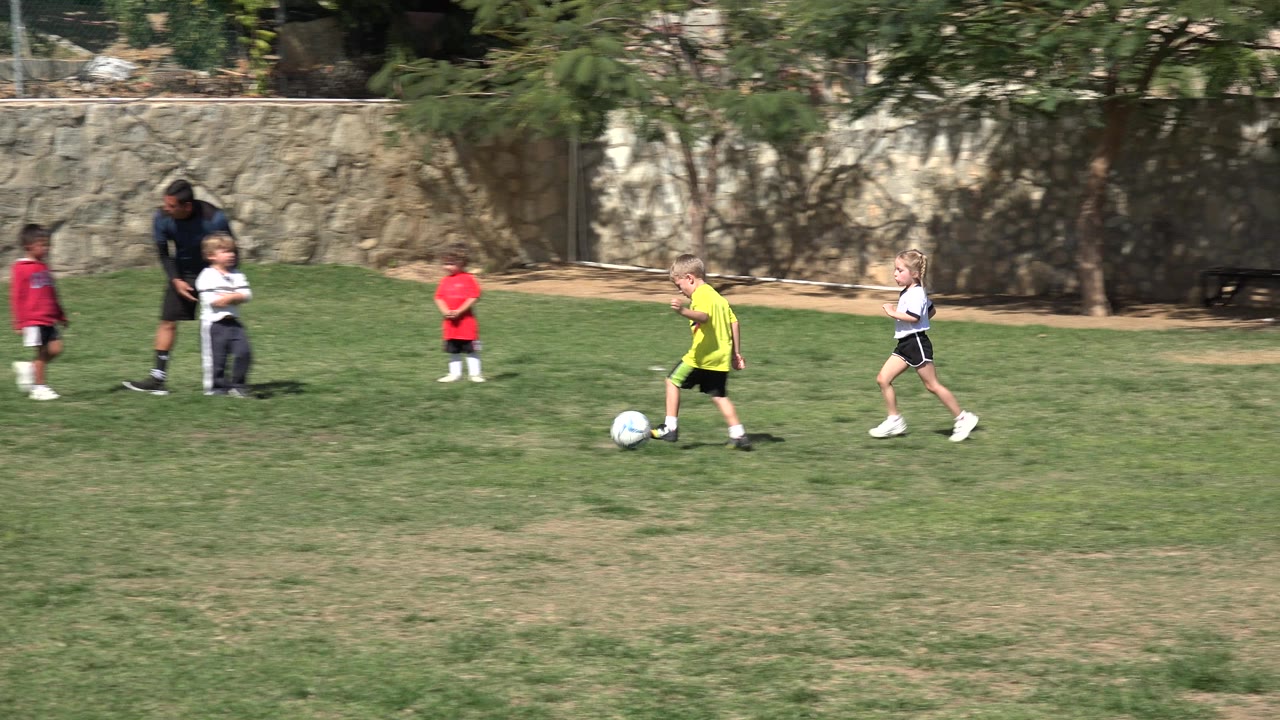A child in a yellow shirt moves the soccer ball across the Los Cabos grass field while other children and the coach remain spread across the background.
