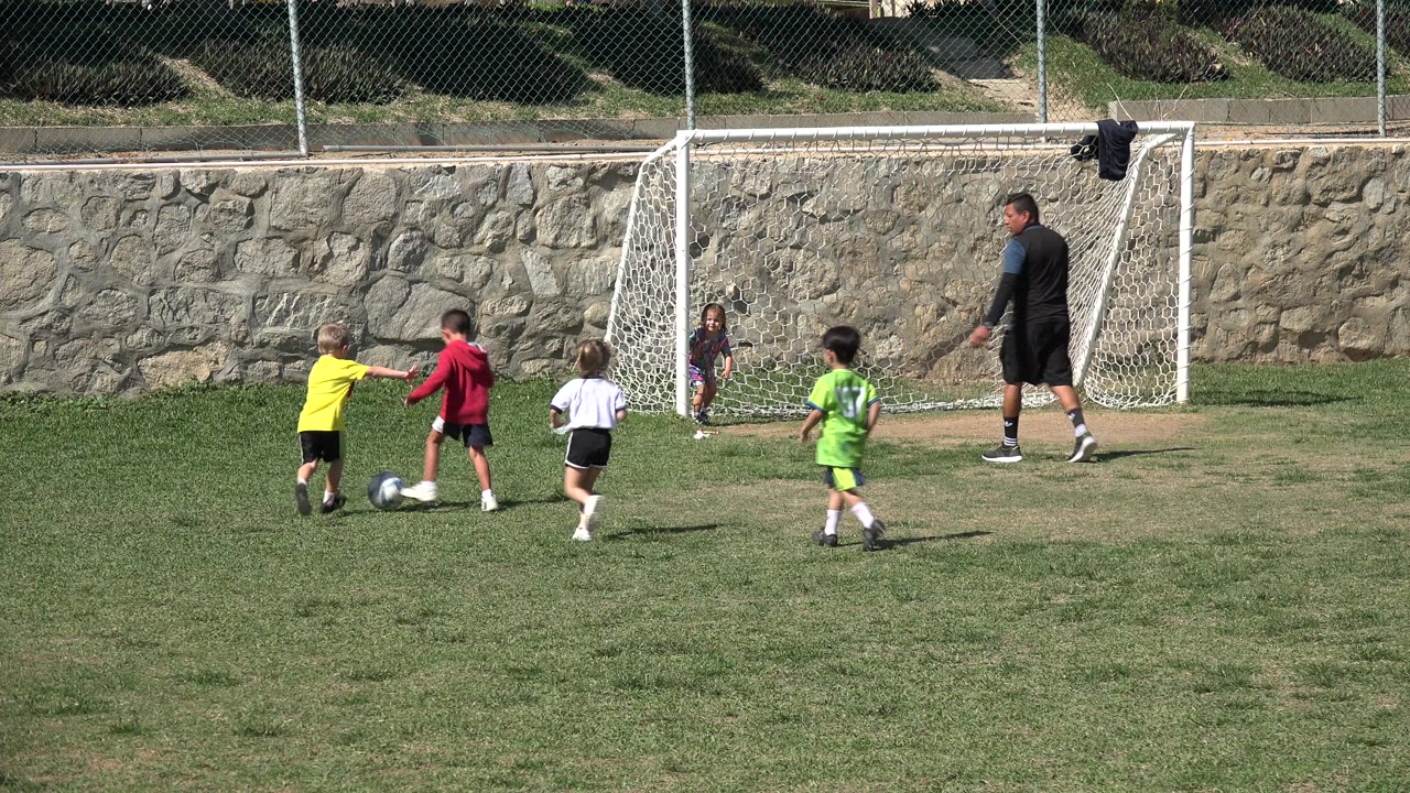 In Los Cabos, a child in a red top is closest to the soccer ball while other children run toward the small goal and the coach watches from the right.