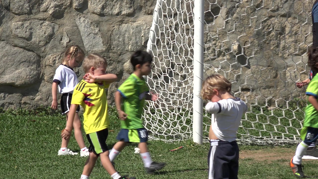 At 0:01 in Los Cabos, Mexico, children move across the grass as a child in a white shirt stands near the net and a child in yellow crosses the foreground.