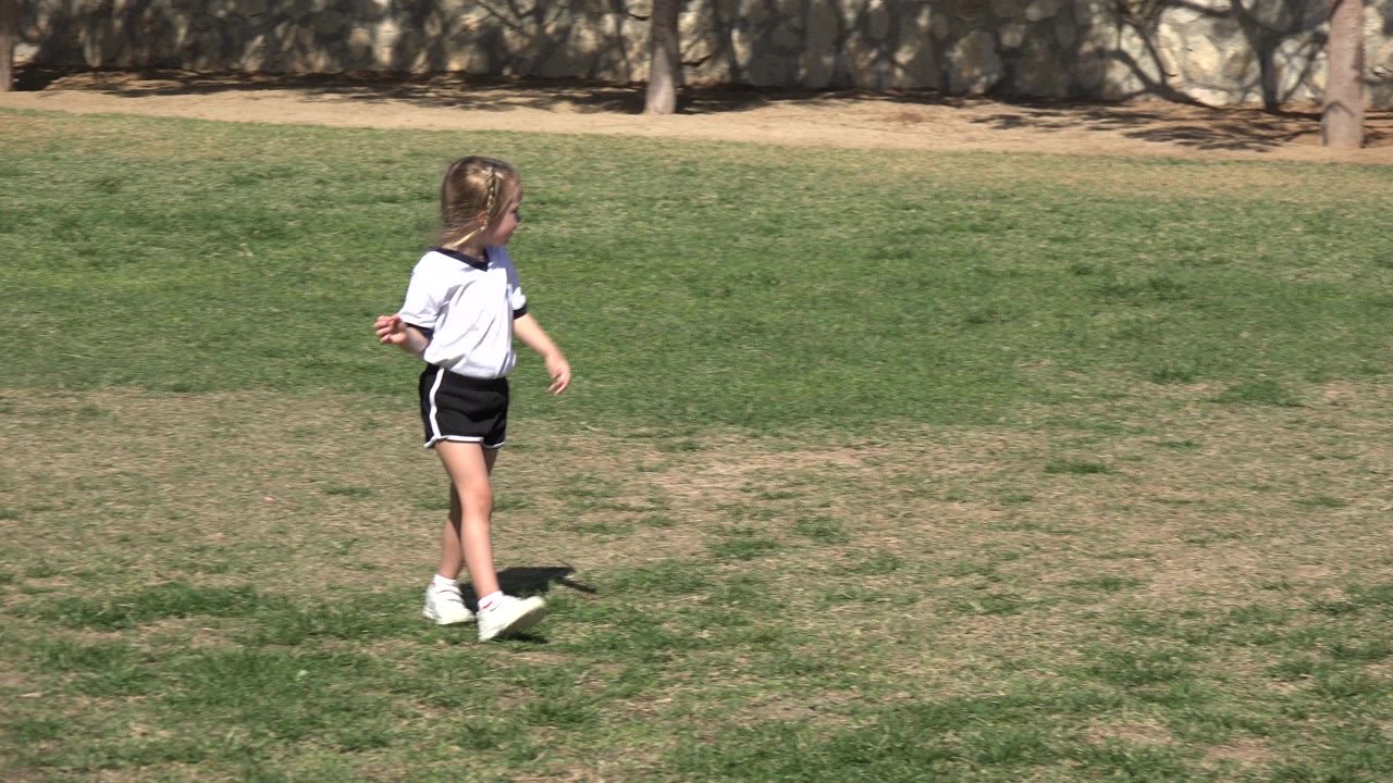 Young girl in a white soccer shirt walking across a grassy field.