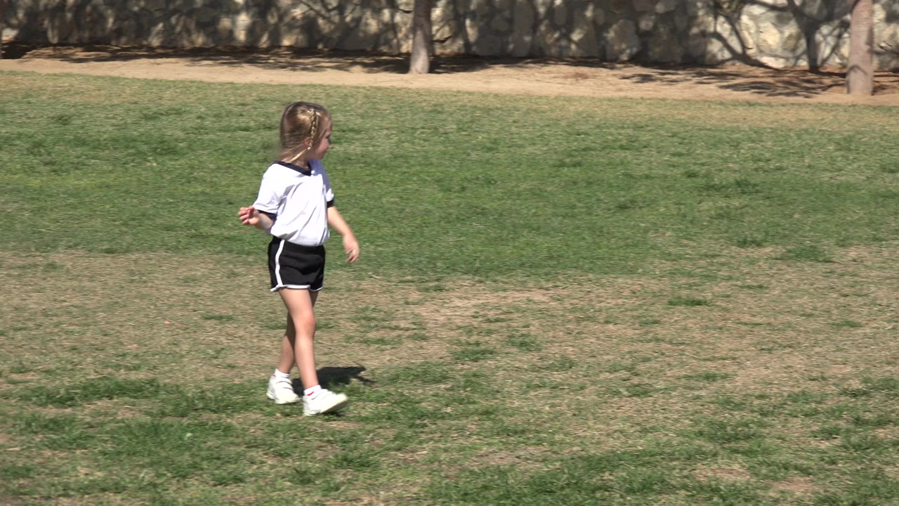 The blonde girl continues across the sunlit grass in Los Cabos, Mexico, looking to the side while the field and stone wall remain in the background.