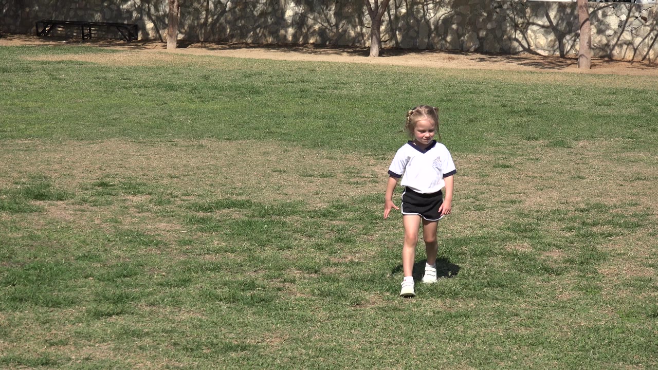 The child remains near the center of the Los Cabos grass field, facing the camera in bright sun with patchy green and brown turf around her.