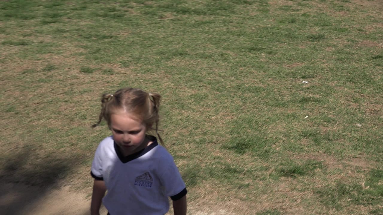 Young girl in a white sports shirt standing near grass and shade.