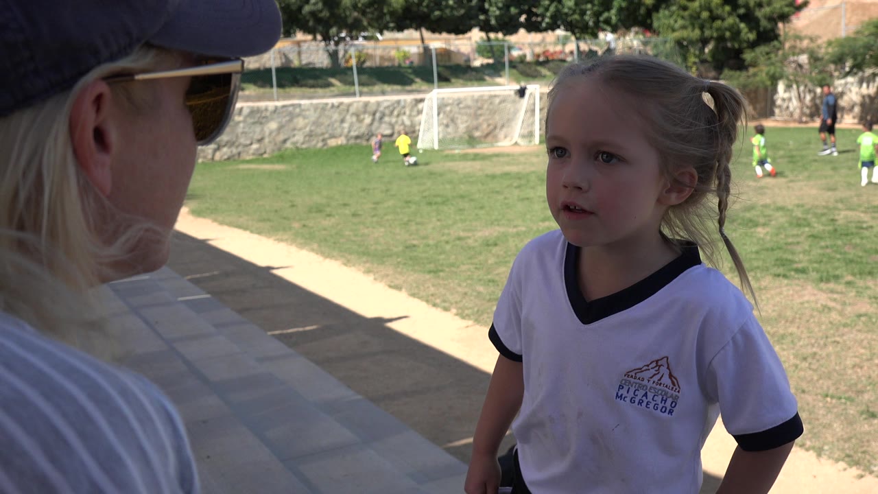 Young girl in a white soccer shirt faces an adult beside a soccer field.