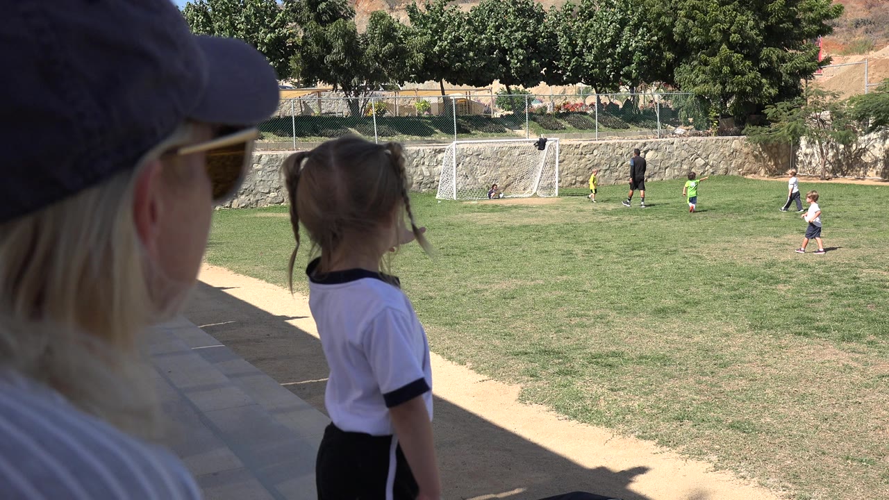 At 0:01 in Los Cabos, Mexico, the young girl remains near the shaded sideline with the adult spectator partly blocking the left side and children playing near the goal in the distance.