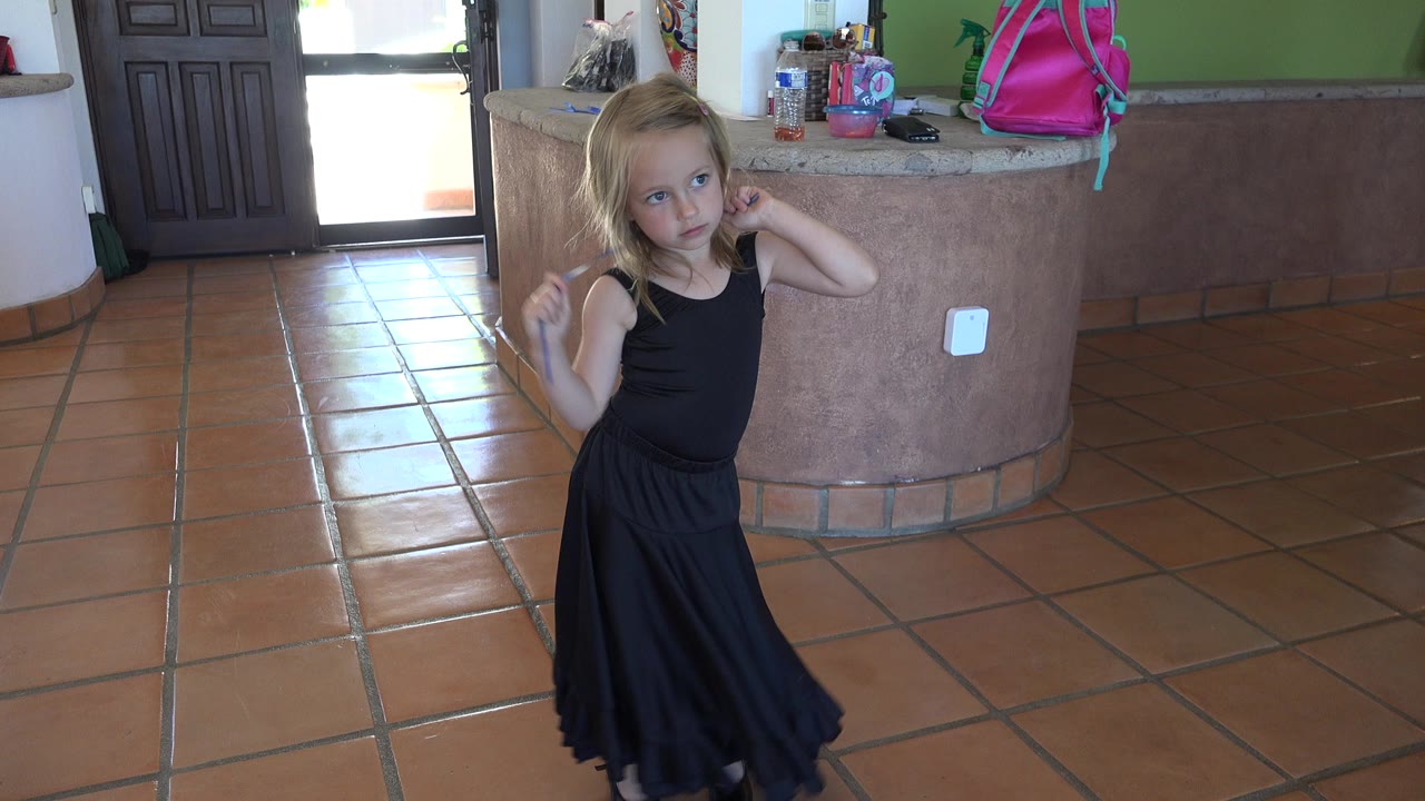 Blonde child in a black dress holding a blue ribbon indoors.