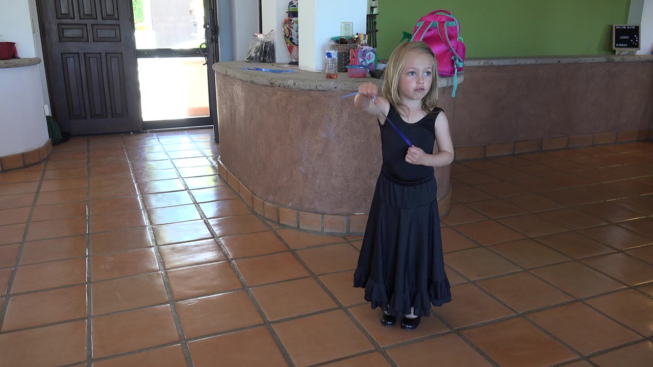 Young girl in a black dress holds a blue ribbon on a terracotta tile floor.