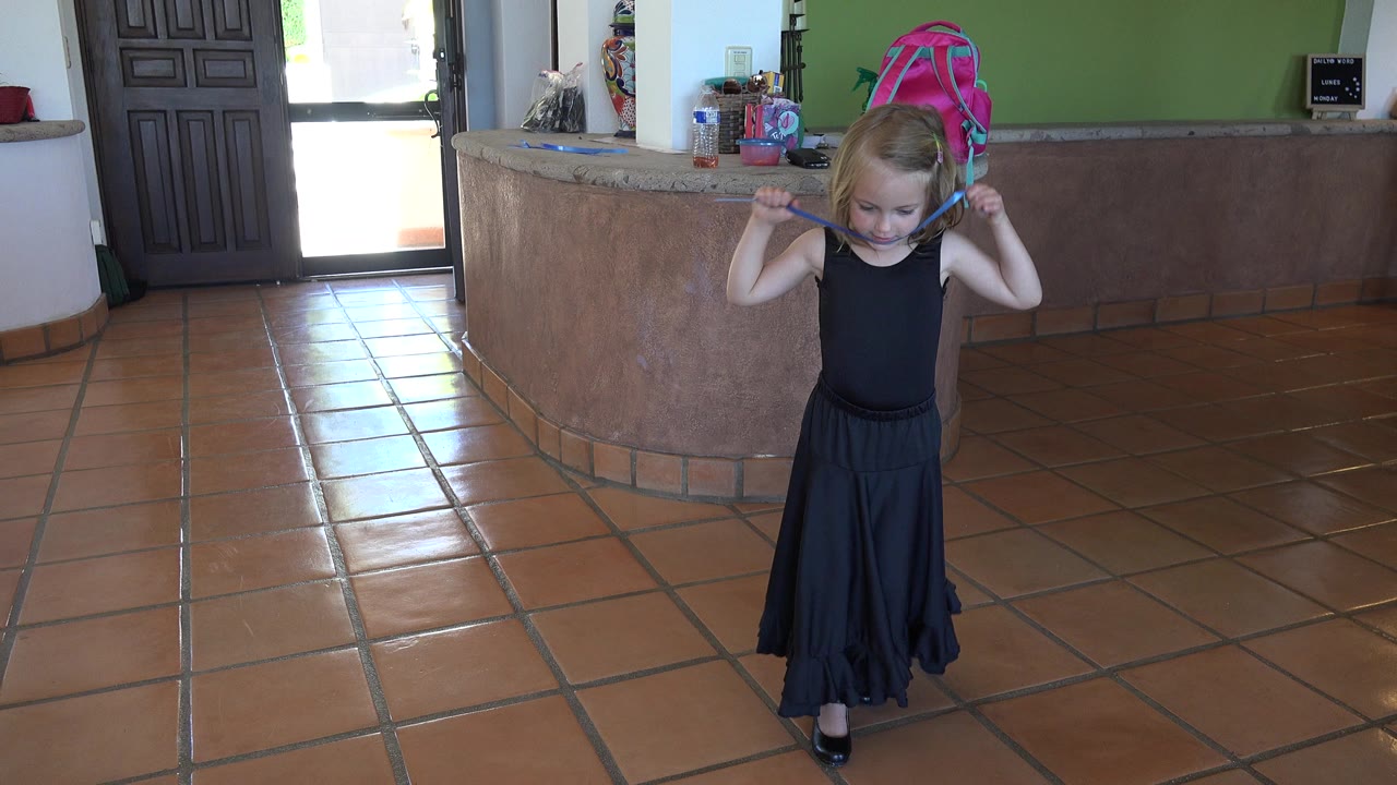A young blonde girl in a black dress stands on terracotta tile in Los Cabos, Mexico, holding a blue object behind her shoulders near a curved counter.