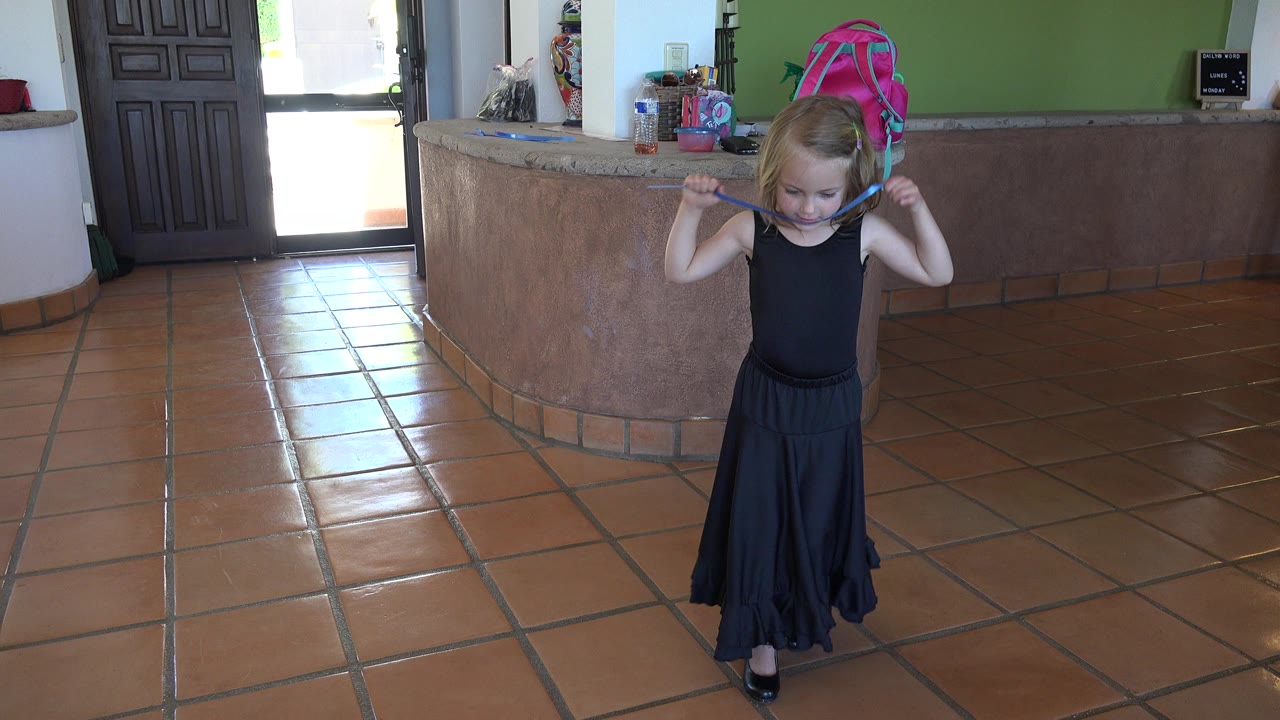 The girl remains near the center of the tiled room in Los Cabos, Mexico, with the blue object looped near her neck and a pink backpack visible on the counter behind her.