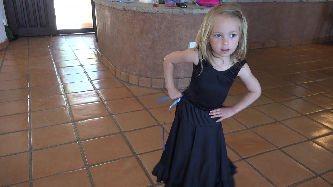 Young blonde girl in a black dress stands with hands on hips on a tiled floor.