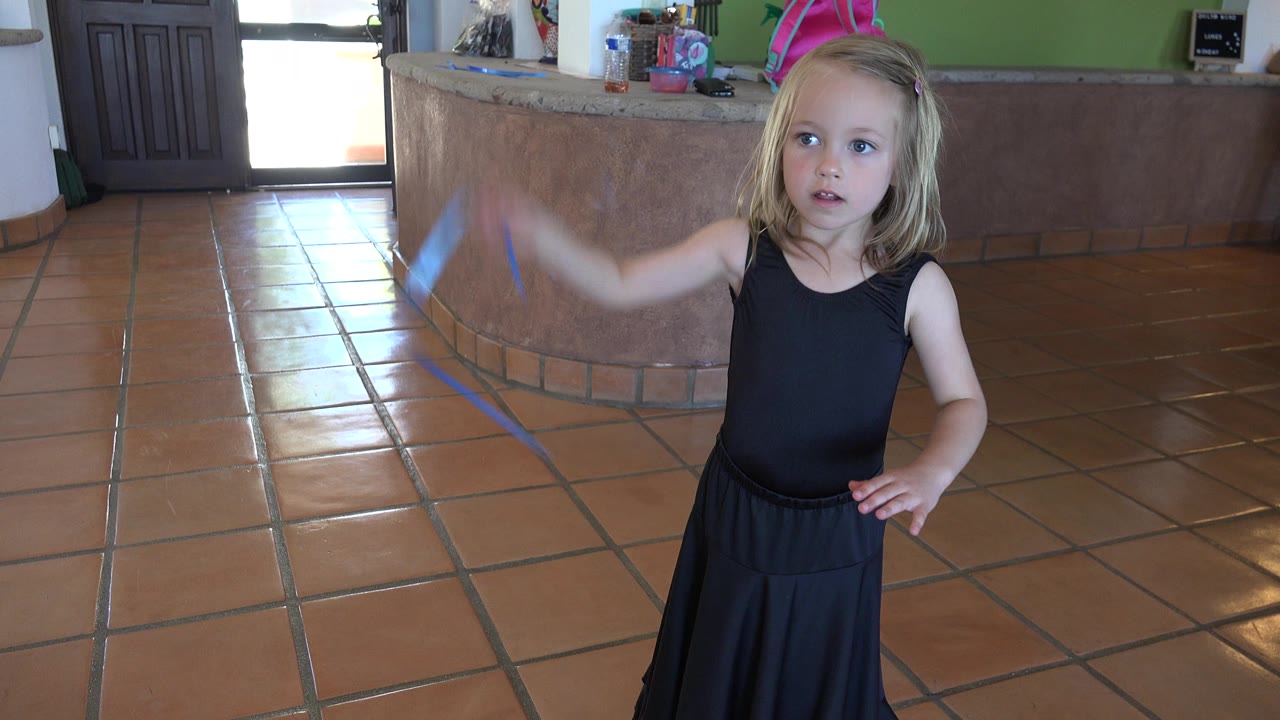 At 0:01 in Los Cabos, Mexico, the girl faces forward with one hand near her waist as the blue ribbon blurs from her raised arm.