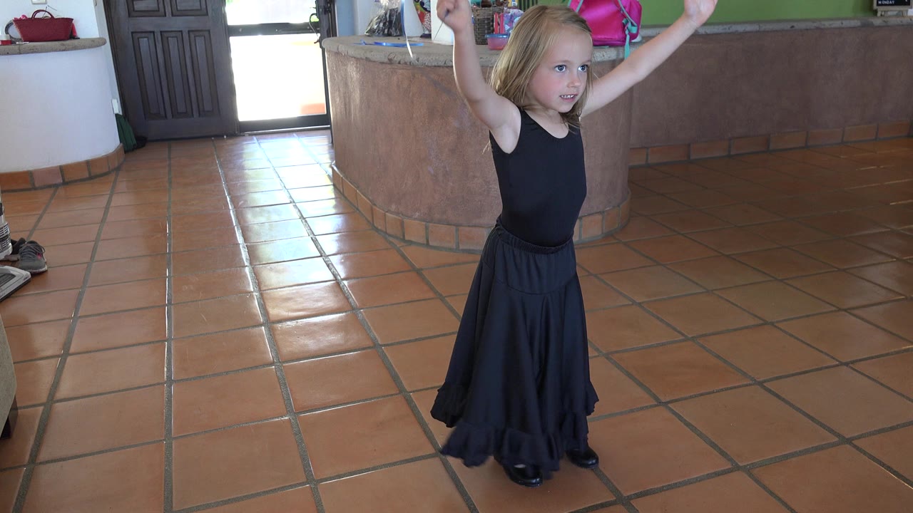 Young girl in a black dress raises both arms on a tile floor.