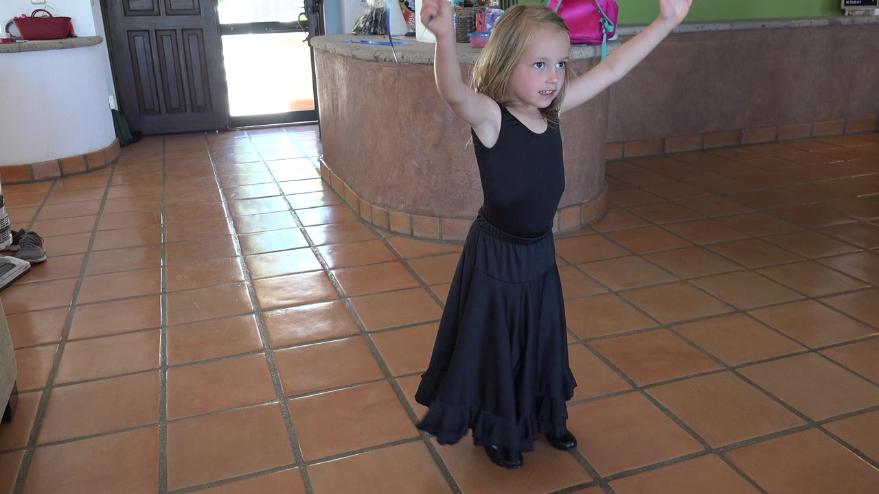 The girl remains in the midground of the Los Cabos room with arms lifted, bright doorway light at left, and a counter behind her.