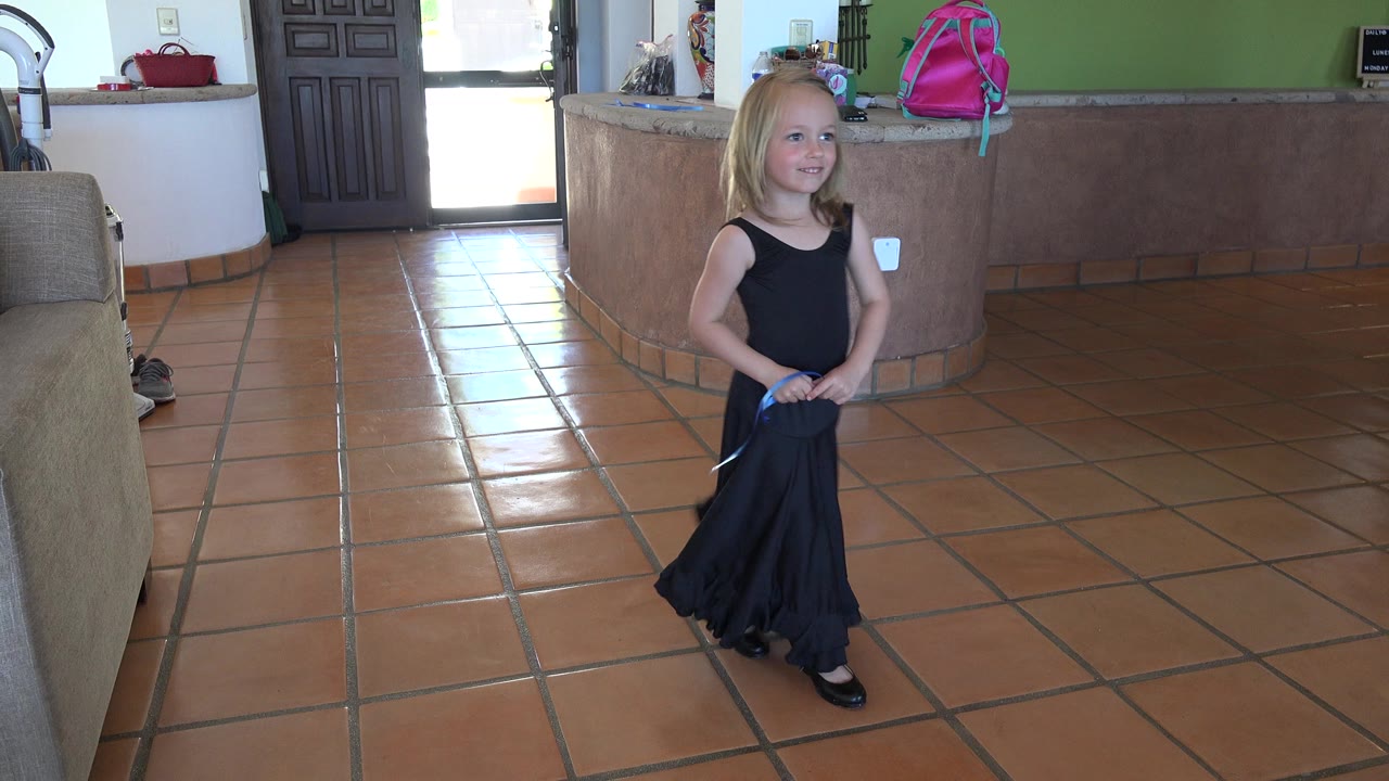 In Los Cabos, Mexico, a blonde little girl in a black dress stands near the center of a tiled room with a couch edge, doorway, counter, and pink backpack behind her.