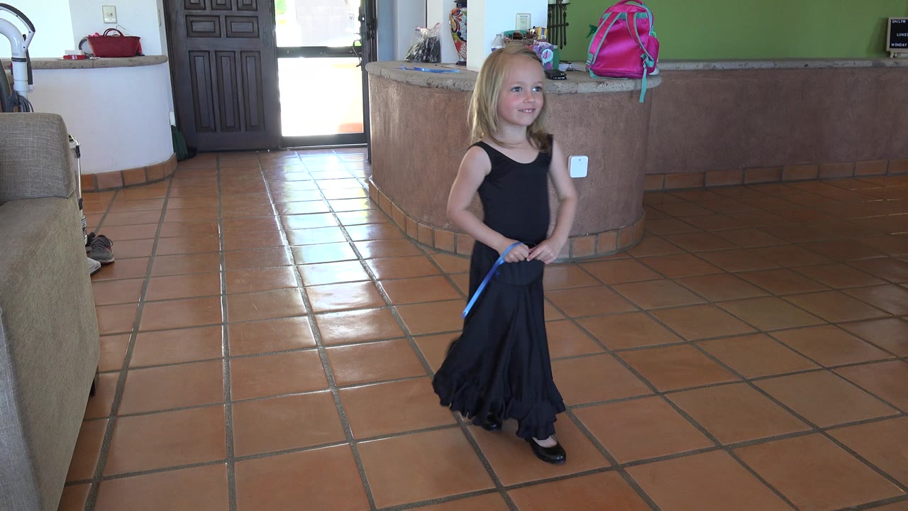 In Los Cabos, Mexico, the girl smiles while holding a blue wand or strap at her waist, with the terracotta floor and kitchen-style counter clearly visible behind her.
