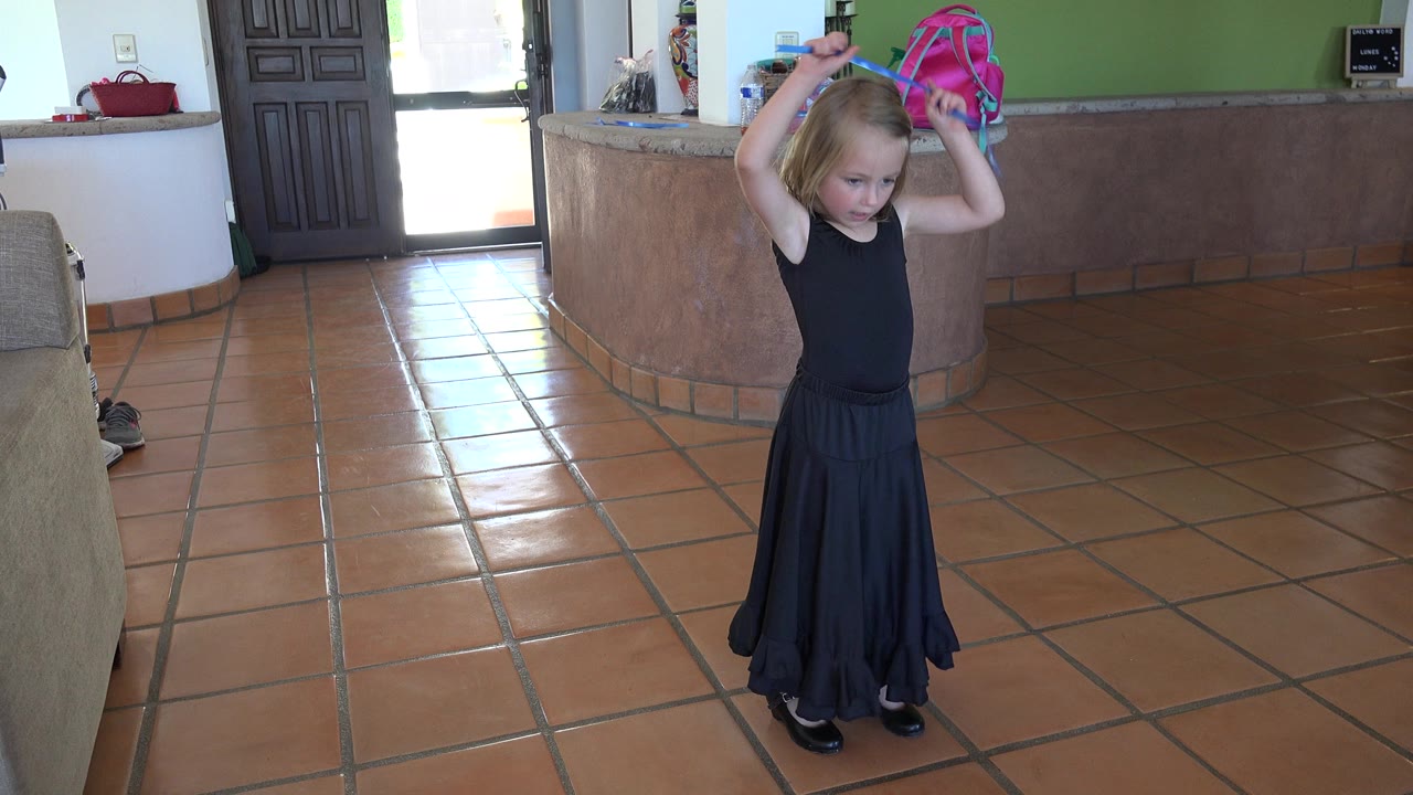 In Los Cabos, Mexico, a young girl in a black dress stands on terracotta tile while raising a blue ribbon above her head near a sofa and curved counter.