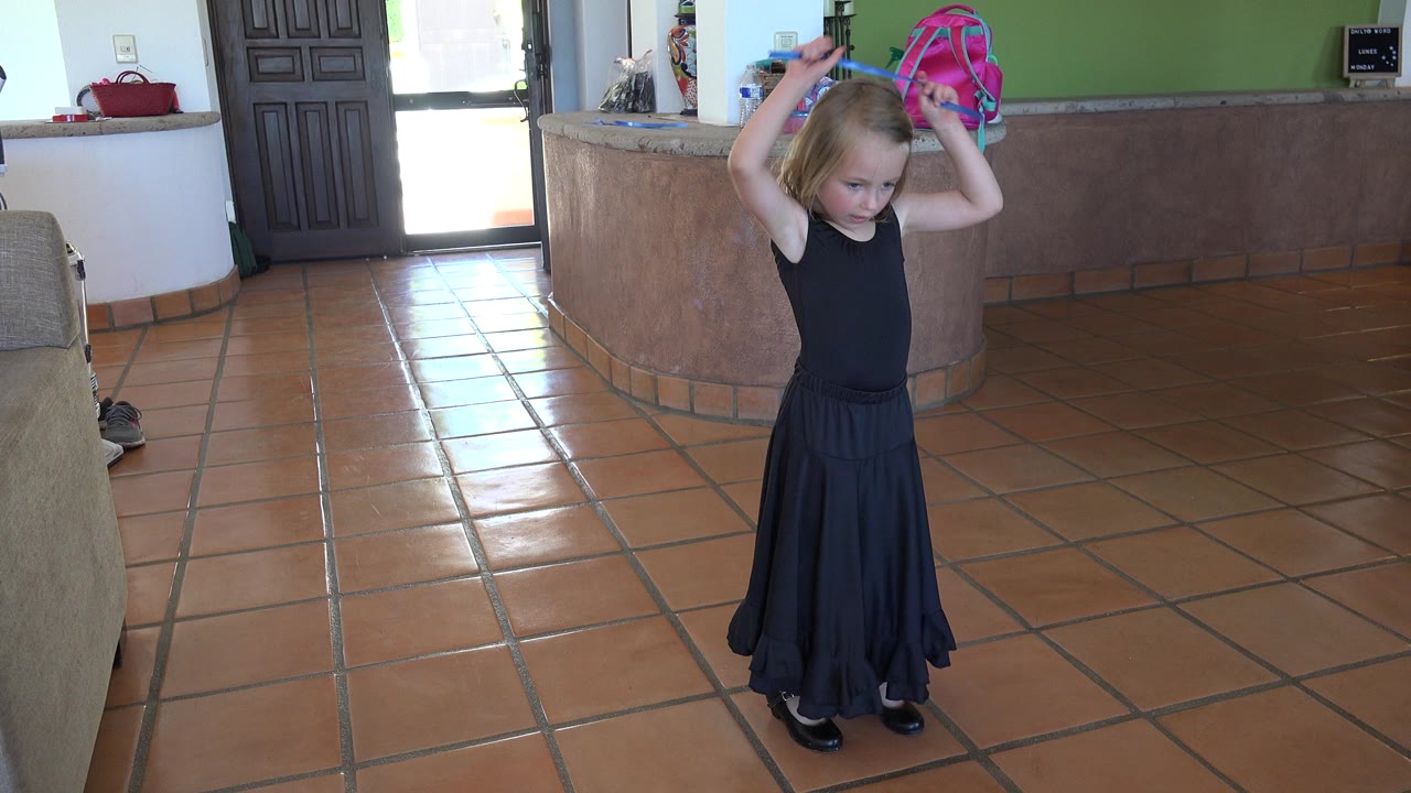 In Los Cabos, Mexico, the girl keeps the blue ribbon raised over her head while standing full body on the tiled floor with daylight near the door behind her.