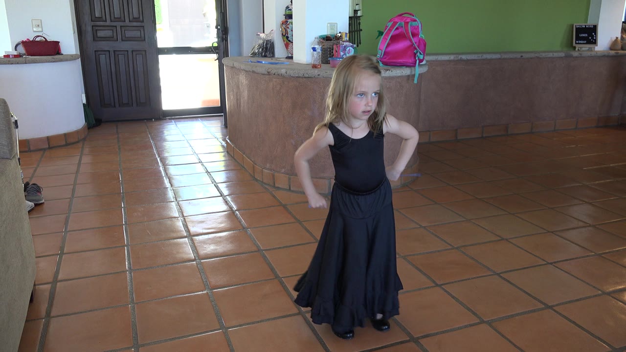 Young child in a black dress standing on a tiled kitchen floor.