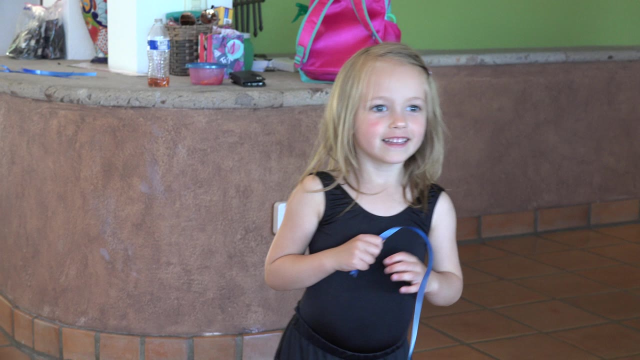 The girl smiles slightly while gripping the blue rope, with the curved counter, tiled floor, and household items visible in the Los Cabos interior.