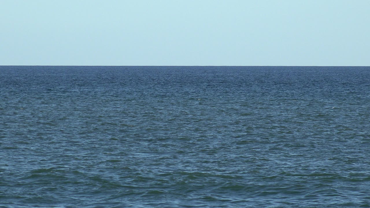 Wide Los Cabos ocean view with rippled blue-green water, pale blue sky, a level horizon, and no visible people at the start of the clip.