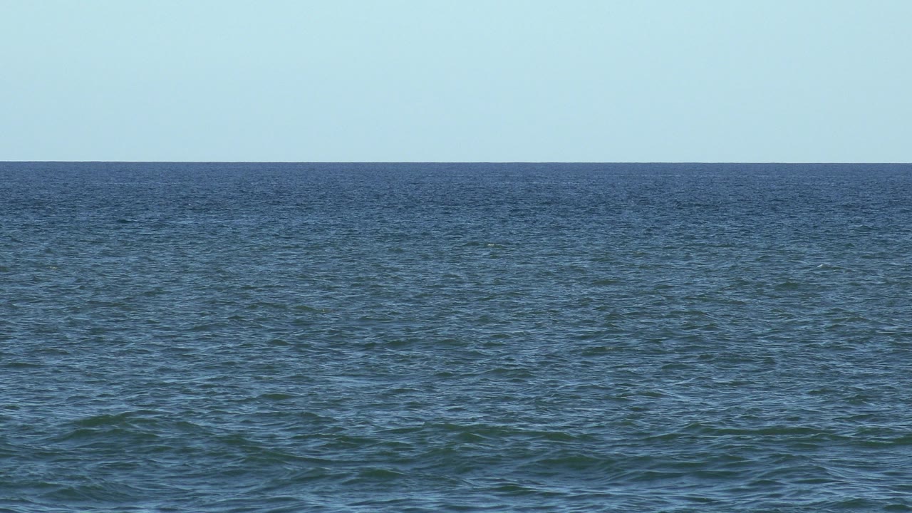 Los Cabos sea fills the frame with a straight horizon, calm surface texture, pale sky, and no visible people one second into the clip.