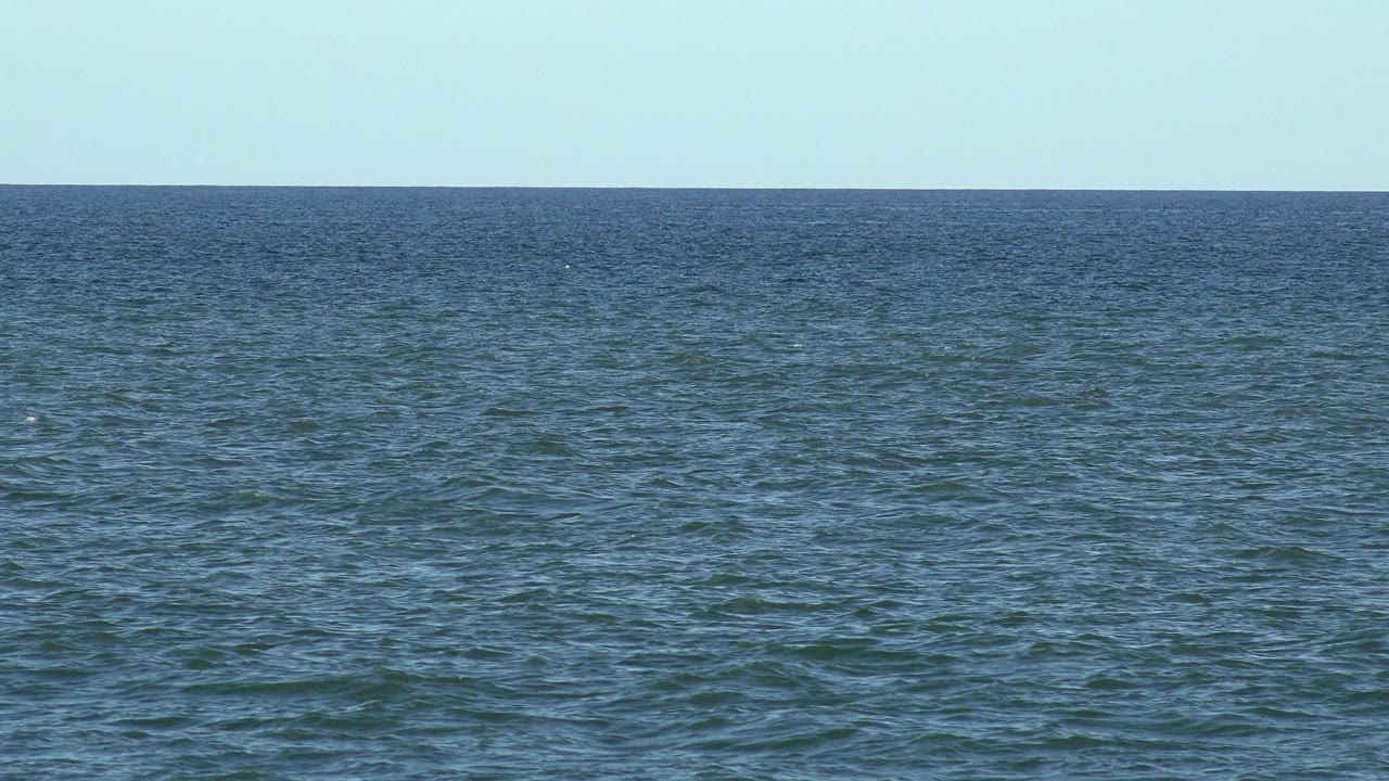 Los Cabos ocean surface at 0:01 with darker rippled water in the foreground, a narrow distant horizon, pale sky, and no visible people.