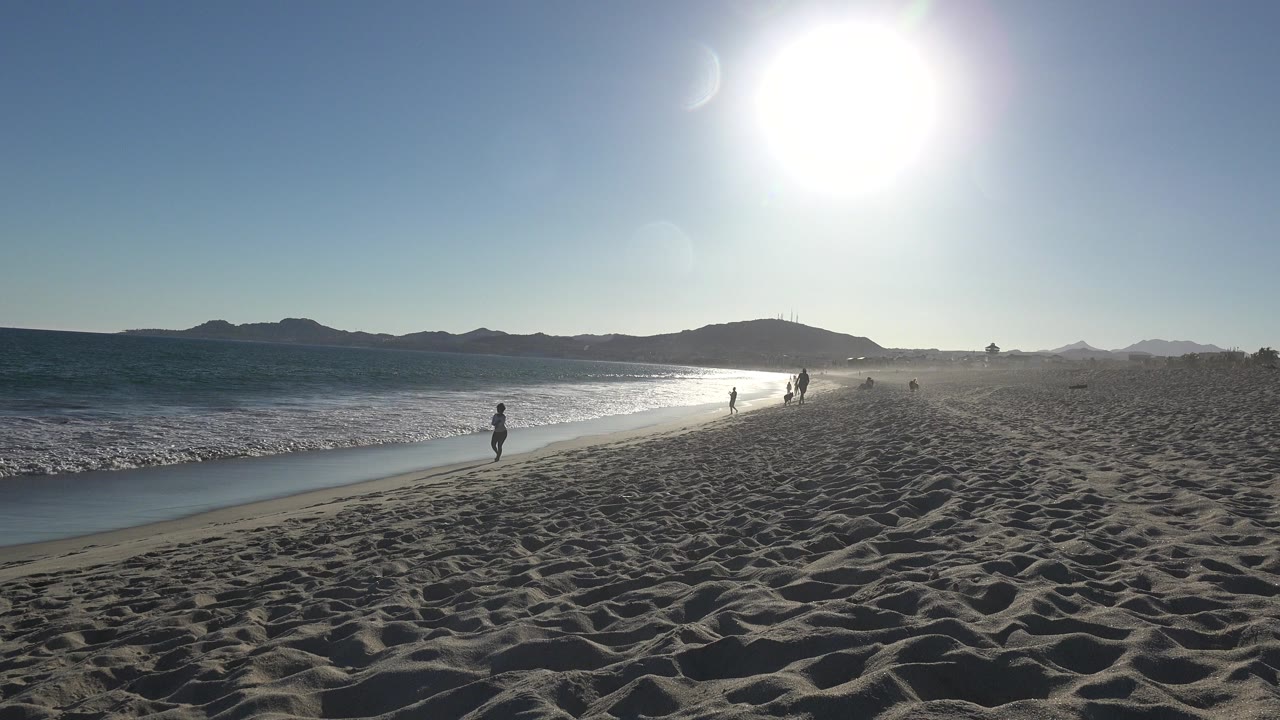 Backlit beach with walkers near the shoreline and low sun overhead.