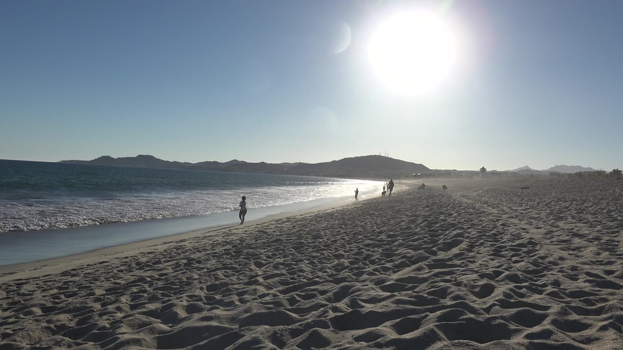 Los Cabos shoreline at 0:01 showing a lone bather nearer the surf, more small figures ahead, bright glare on the water, and dark hills beyond.