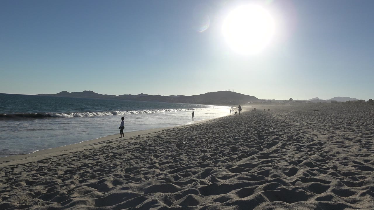 Sunny beach with waves, sand, distant mountains, and people near the shoreline.