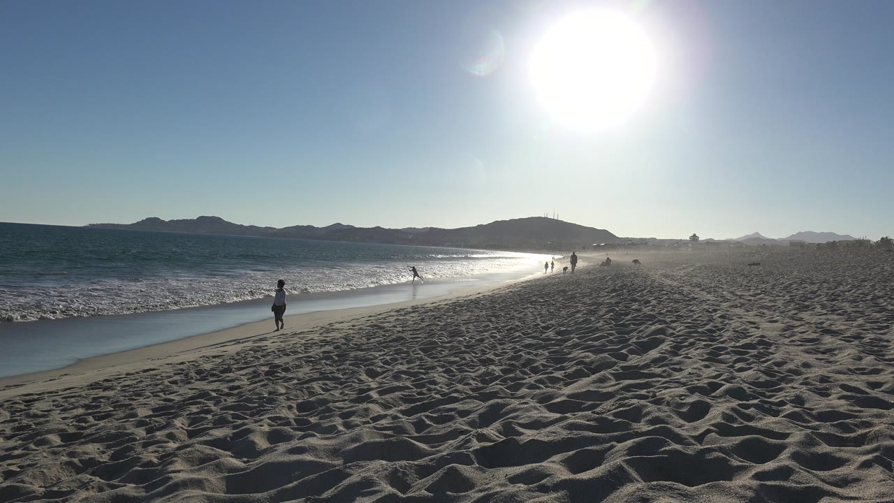 Wide sunny beach with surf, sand, distant hills, and small people on the shoreline.